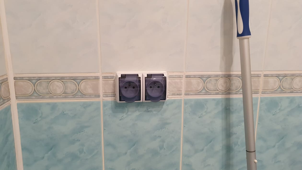 Dwa gniazdka elektryczne z klapkami ochronnymi zamontowane na ścianie z błękitnych płytek, widoczny fragment mopa.