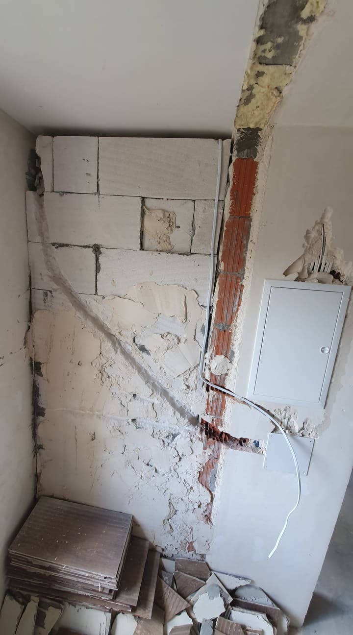 Ściana w trakcie prac murarskich z widocznymi przewodami elektrycznymi i skrzynką rozdzielczą, częściowo otynkowana, z ułożonymi płytkami pod ścianą.