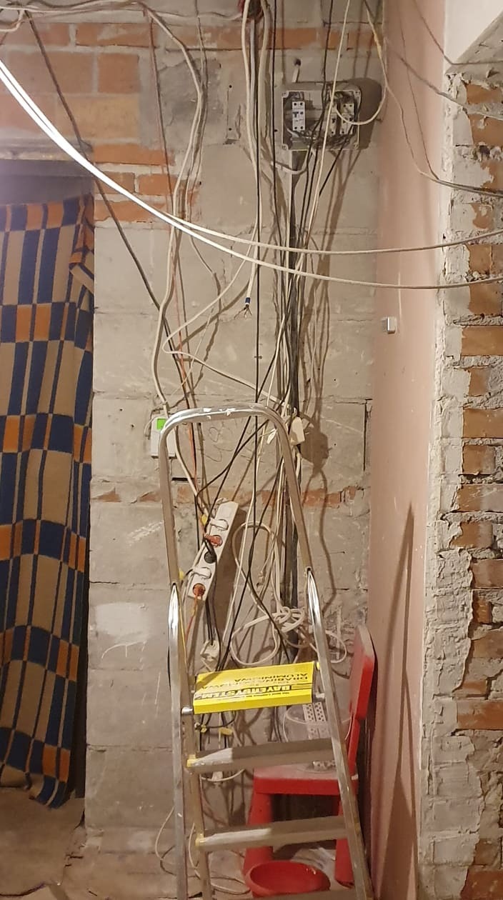Ściana w trakcie remontu z odsłoniętą instalacją elektryczną, widoczne kable, puszki elektryczne, drabina aluminiowa z żółtą naklejką i fragment zasłony w kratę.