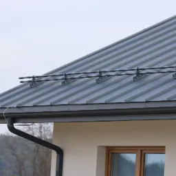 Szary dach z blachy trapezowej z zamontowanymi zabezpieczeniami przeciwśniegowymi i orynnowaniem w kolorze grafitowym, widok z dołu na tle drzew.