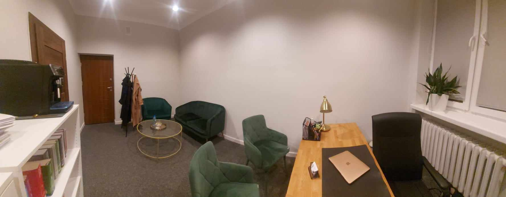 Wnętrze biura w Lublinie z zielonymi fotelami, drewnianym biurkiem z laptopem i złotą lampką, oraz rośliną doniczkową na parapecie.