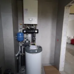 Nowoczesna kotłownia z piecem gazowym Immergas, zbiornikiem, niebieskim naczyniem przeponowym i rozbudowaną instalacją hydrauliczną w pomieszczeniu wykończonym szarą płytką.