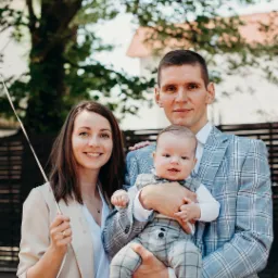 Uśmiechnięta rodzina: mama z balonem, tata w niebieskiej marynarce trzymający ubranego w szary garnitur synka na tle zieleni