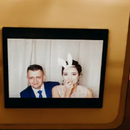 Zdjęcie pary w fotobudce: mężczyzna w niebieskim garniturze i kobieta z papierowymi uszami królika, zasłaniająca usta palcem.