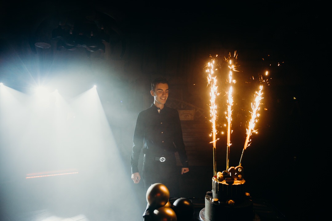 Młody mężczyzna w czarnej koszuli i spodniach, uśmiechnięty, stoi obok tortu urodzinowego z fontannami iskier w ciemnym otoczeniu z efektem reflektorów.