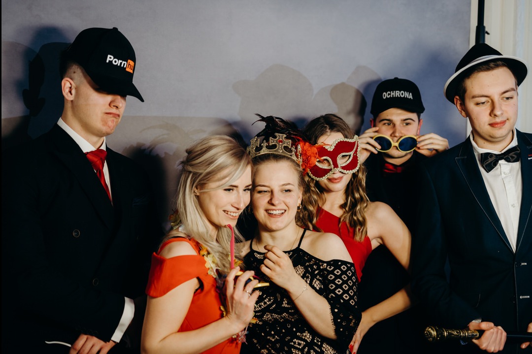 Grupa uśmiechniętych młodych osób pozuje do zdjęcia w fotobudce, ubrani w eleganckie stroje i śmieszne rekwizyty, takie jak maski, korony i zabawne okulary, na tle wzorzystej ściany.