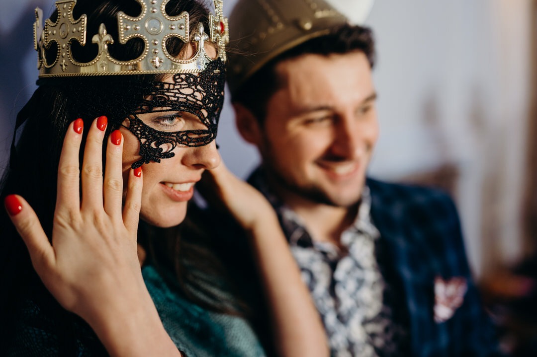 Uśmiechnięta kobieta w koronie i czarnej koronkowej masce, z czerwonym manicurem, pozuje z uśmiechniętym mężczyzną w papierowej koronie.