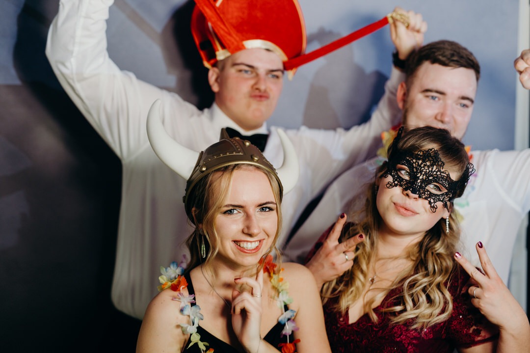 Grupa czterech osób pozuje w przebraniach: kobieta w hełmie wikinga, kobieta w czarnej koronkowej masce, mężczyzna w czerwonym kapeluszu karnawałowym i mężczyzna z kwiatami na szyi, wszyscy...