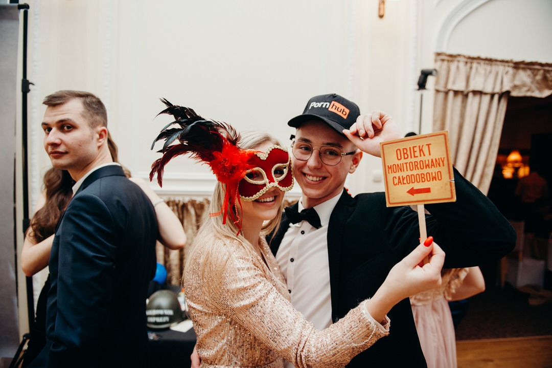 Para w przebraniach pozuje z rekwizytami podczas eventu z fotobudką: kobieta w złotej sukience i masce z piórami, mężczyzna w smokingu i czapce z napisem, trzymający tabliczkę z napisem 'Obiekt...