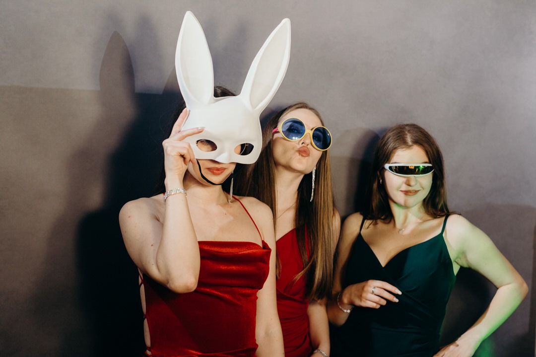 Trzy kobiety w wieczorowych sukienkach pozują z rekwizytami: maską królika i futurystycznymi okularami przed szarym tłem fotobudki.