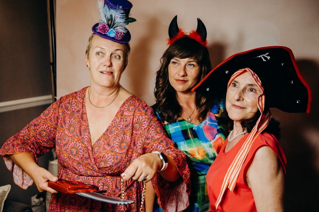 Trzy kobiety w przebraniach: jedna w mini-kapeluszu z piórami, druga w opasce z rogami, trzecia w pirackim kapeluszu, pozują z rekwizytami w postaci zabawkowego noża trzymanego przez kobietę...