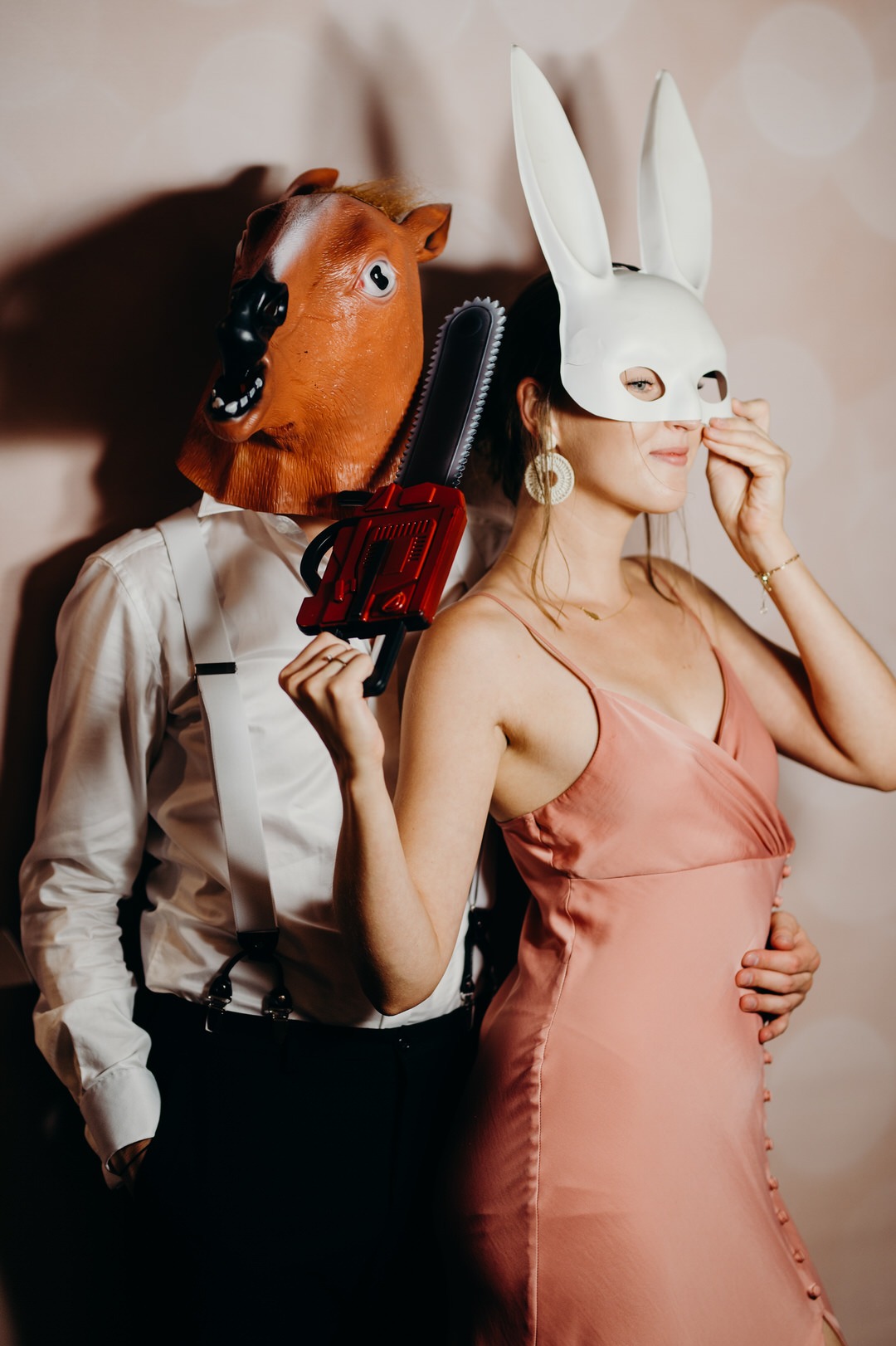 Para w maskach: mężczyzna w masce konia trzyma czerwoną zabawkowa piłę łańcuchową, kobieta w białej masce królika poprawia maskę, oboje w eleganckich strojach na tle jasnego tła.