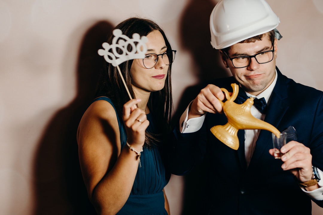 Para pozuje z rekwizytami: kobieta z koroną na patyku, mężczyzna w kasku budowlanym wlewa coś z lampy Alladyna do kieliszka.