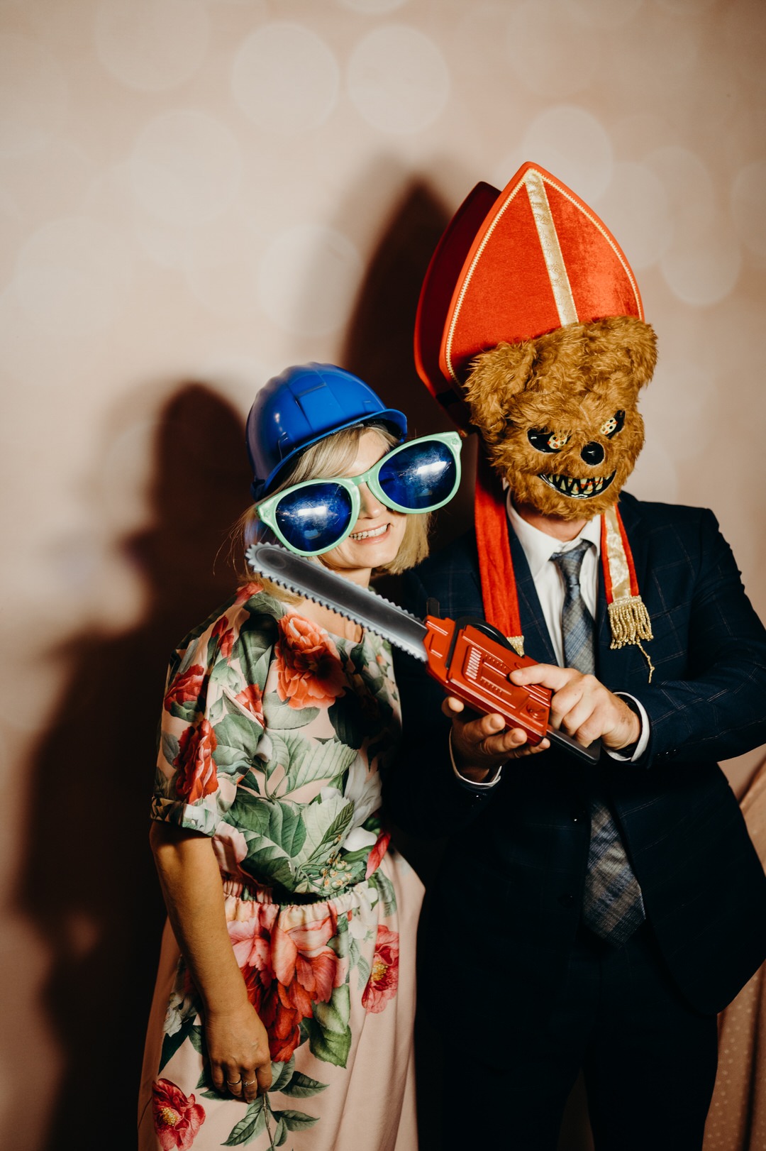 Para w przebraniach pozuje z rekwizytami: kobieta w kasku i okularach, mężczyzna w masce misia i stroju biskupa trzyma zabawkowa piłę łańcuchową.