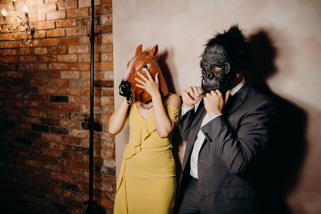 Dwie osoby w maskach: kobieta w masce konia i żółtej sukience zasłaniająca twarz dłońmi, mężczyzna w masce goryla w garniturze, pozujący na tle ściany z cegły i oświetlenia.