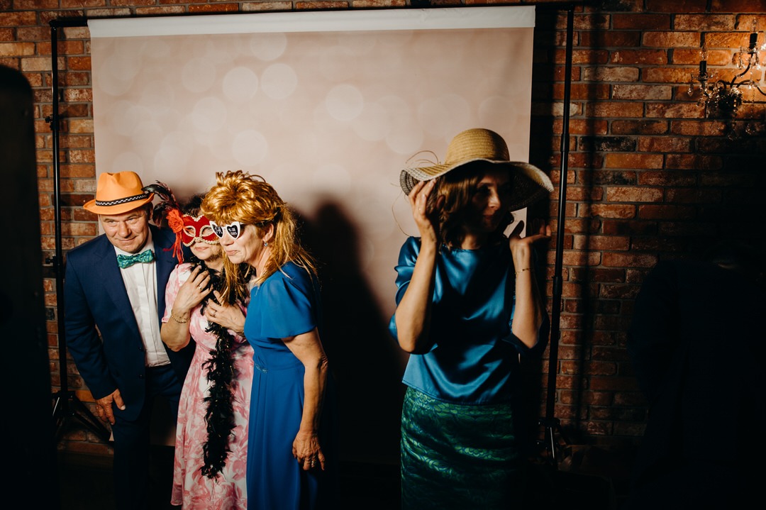 Grupa osób pozuje w fotobudce z rekwizytami: kapelusze, maski i boa, ściana z cegły w tle.