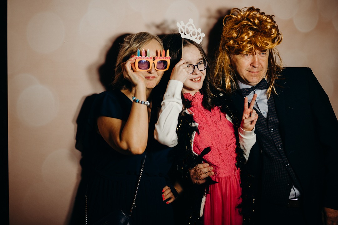 Trzy osoby pozują w fotobudce z rekwizytami: kobieta w okularach z płomieniami świeczek, dziewczynka z papierową koroną na patyku i mężczyzna w peruce z długimi blond włosami, pokazujący znak...