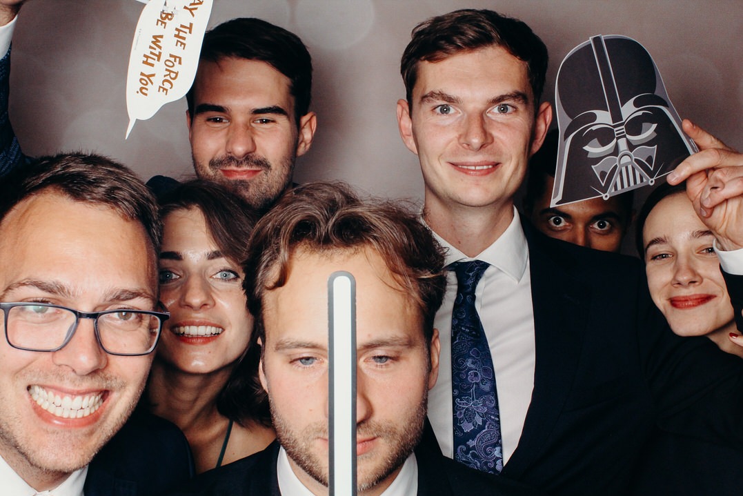 Grupa uśmiechniętych osób pozuje do zdjęcia z rekwizytami, w tym maską Dartha Vadera i dymkiem z napisem 'May the Force be with you'.
