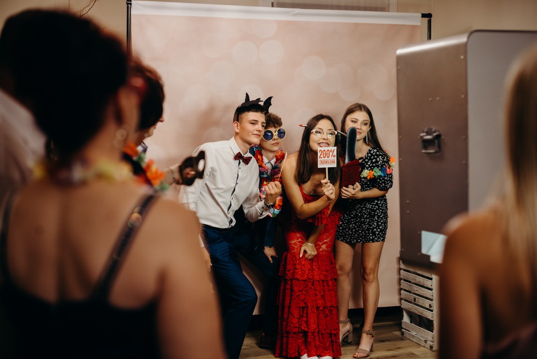 Grupa rozbawionych osób pozuje z rekwizytami przed tłem i automatem do zdjęć, trzymając tabliczkę z napisem '200% zadowolenia', jeden z uczestników ma rogi diabła na głowie.