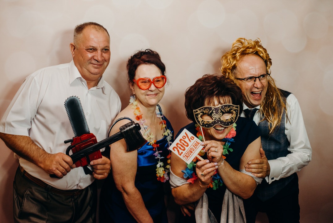 Cztery osoby pozują z rekwizytami: plastikową piłą łańcuchową, maską karnawałową, okularami w kształcie serc i peruką podczas zabawy w fotobudce.