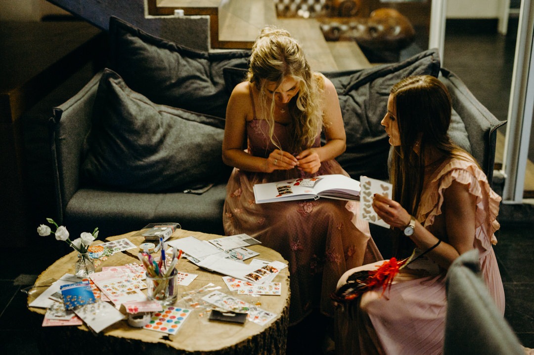 Dwie kobiety w sukienkach siedzą przy okrągłym stoliku z pnia drzewa, ozdabiają album naklejkami i zdjęciami z fotobudki.