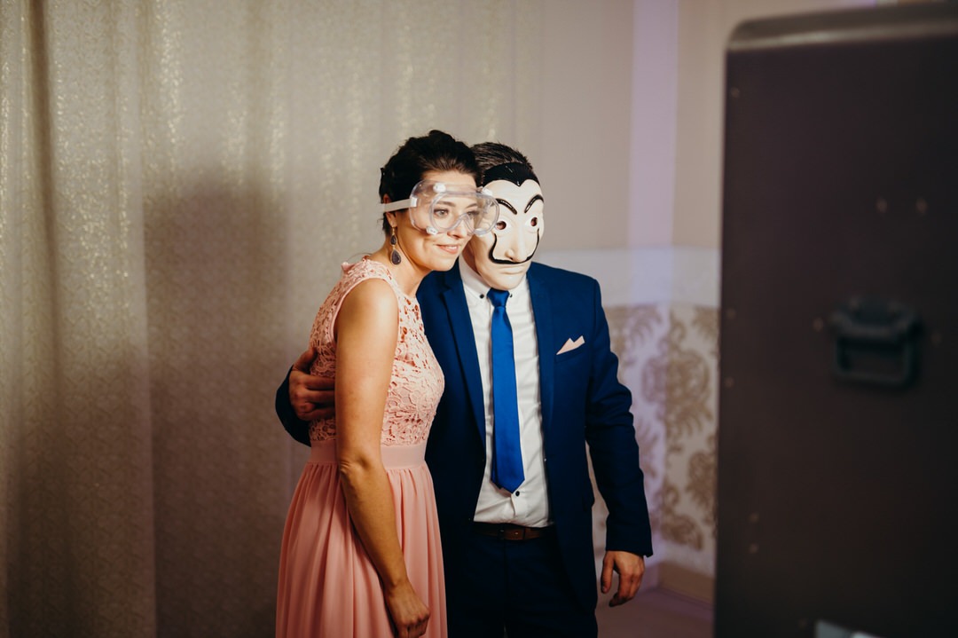 Para w przebraniach pozuje do zdjęcia w fotobudce: kobieta w różowej sukience i okularach ochronnych, mężczyzna w niebieskim garniturze i masce Salvadora Dali.