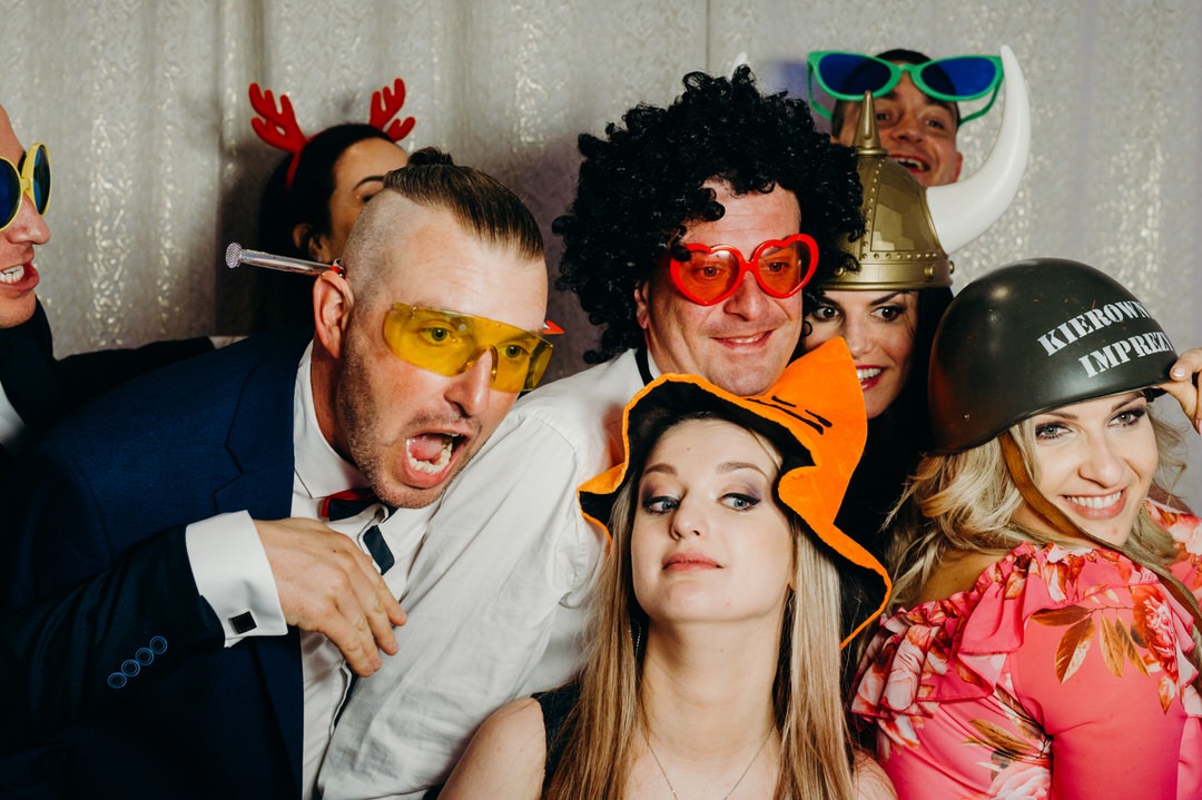 Grupa uśmiechniętych osób w przebraniach pozuje do zdjęcia w fotobudce, widoczne okulary w kształcie serc, hełm z napisem 'Kierownik imprezy', peruka afro i rekwizyty imprezowe.