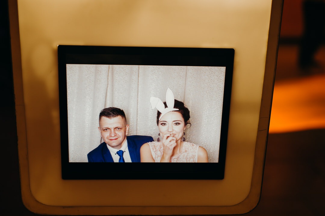 Zdjęcie pary w fotobudce: mężczyzna w niebieskim garniturze i kobieta z papierowymi uszami królika, zasłaniająca usta palcem.