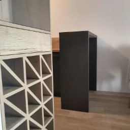 Regał na wino z jasnego drewna z szufladą i szklaną witryną, obok ciemnoszarego blatu na wysokich nogach, na podłodze z paneli drewnopodobnych.