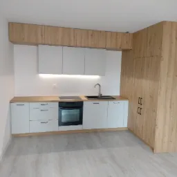 Jasna kuchnia z białymi frontami i drewnianymi akcentami, obejmująca zabudowę pod sufit i szafę w rogu, z czarnym zlewem i płytą grzewczą.