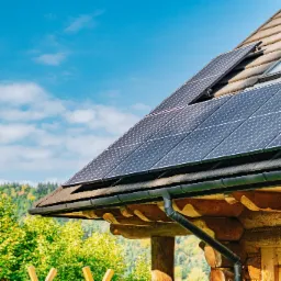 Panele słoneczne na dachu drewnianego domu z oknem dachowym i rynną, w tle drzewa i błękitne niebo z chmurami.