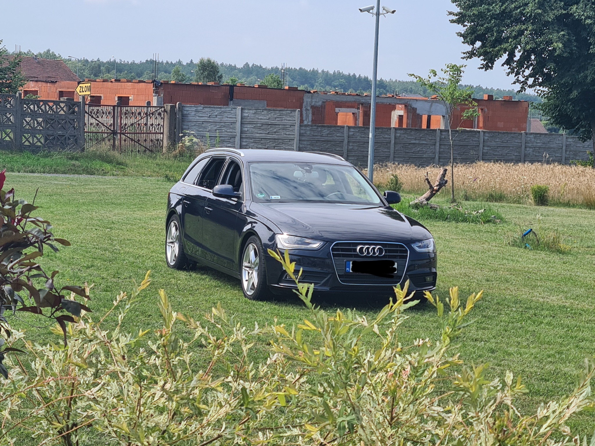 Czarny samochód kombi Audi A4 na trawniku przed budową domu z czerwonej cegły i znakiem 'Złom'.