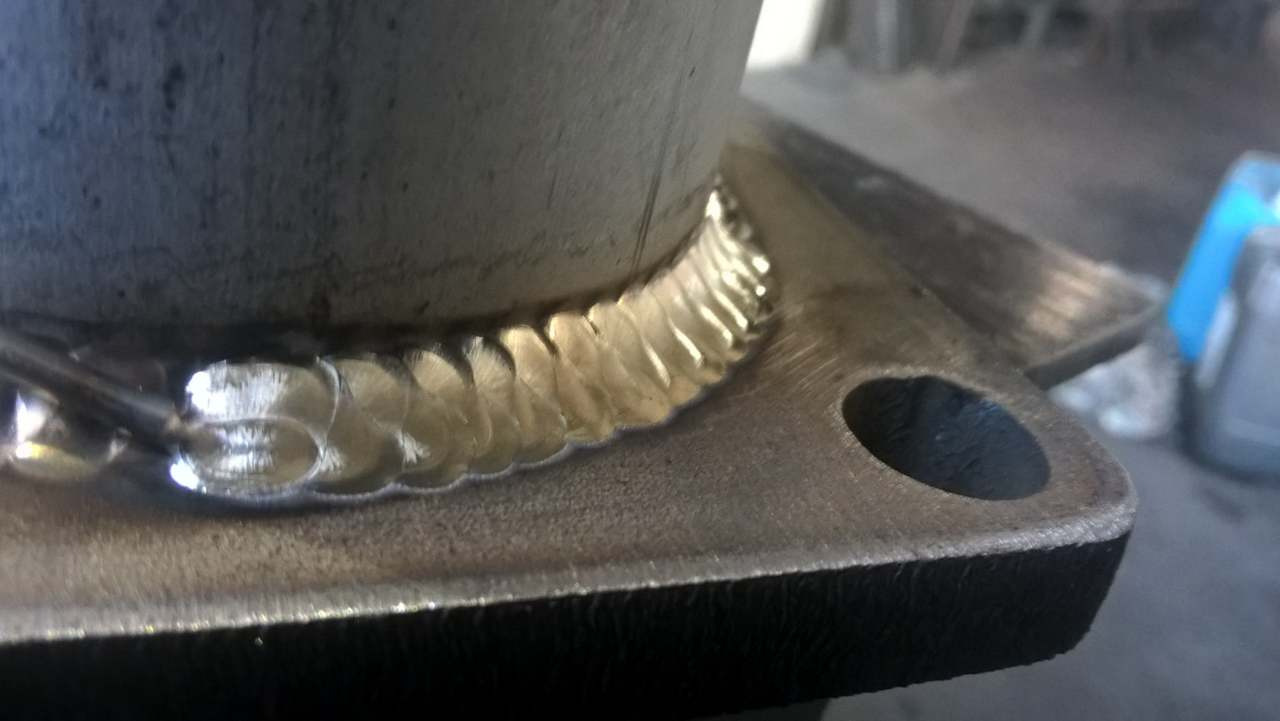 Spoina TIG na stali nierdzewnej, widoczne charakterystyczne łuski spoiny ułożone jedna obok drugiej, łączące rurę z płaskownikiem.