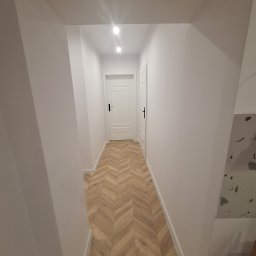 KozFach Oskar Kozik - Korytarz po remoncie z ułożonymi panelami podłogowymi w jodełkę, pomalowanymi na biało ścianami i dwiema białymi drzwiami z czarnymi klamkami.