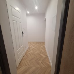 KozFach Oskar Kozik - Jasne wnętrze przedpokoju z nową podłogą ułożoną w jodełkę, białe ściany i drzwi, widoczne oświetlenie sufitowe punktowe.