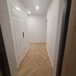 Jasne wnętrze przedpokoju z nową podłogą ułożoną w jodełkę, białe ściany i drzwi, widoczne oświetlenie sufitowe punktowe.