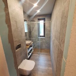 KozFach Oskar Kozik - Nowoczesna łazienka w trakcie remontu, widoczne szare płytki imitujące beton, drewnopodobne panele na podłodze, biała podwieszana toaleta, umywalka nablatowa z drewnianym blatem, szklana kabina...