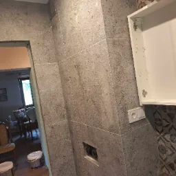 Ściana łazienki wyłożona szarymi, kwadratowymi płytkami imitującymi kamień, z widoczną białą szafką bez frontu, gniazdkiem elektrycznym i puszką instalacyjną. W tle widoczne wiaderka z zaprawą...