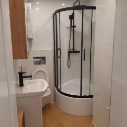 Nowoczesna łazienka z kabiną prysznicową z czarnymi profilami, białą umywalką z czarną armaturą, drewnianą szafką i białymi płytkami na ścianach oraz panelami na podłodze.