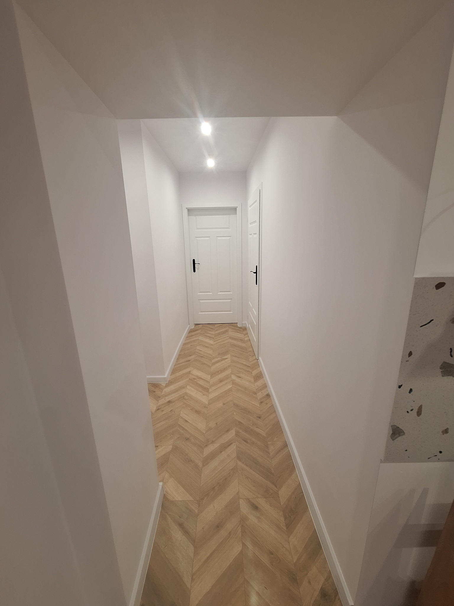 Korytarz po remoncie z ułożonymi panelami podłogowymi w jodełkę, pomalowanymi na biało ścianami i dwiema białymi drzwiami z czarnymi klamkami.