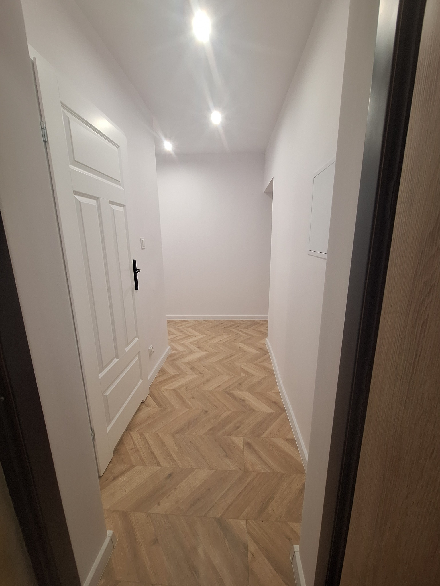 Jasne wnętrze przedpokoju z nową podłogą ułożoną w jodełkę, białe ściany i drzwi, widoczne oświetlenie sufitowe punktowe.
