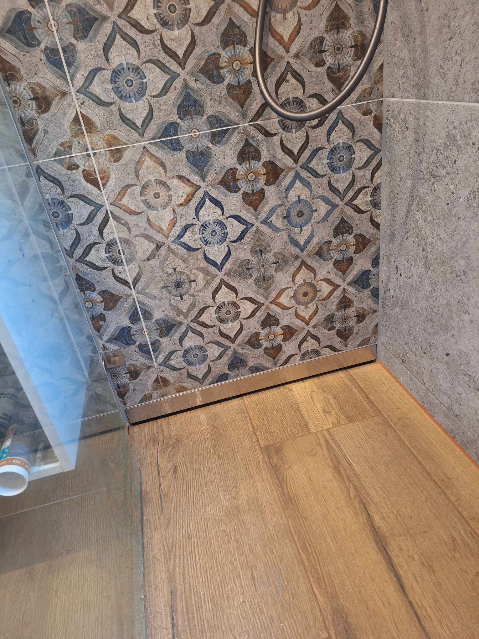 Fragment wykończonej łazienki: połączenie drewnopodobnych paneli podłogowych, ściany z płytek w kwiatowy wzór i szarej ściany, widoczny fragment kabiny prysznicowej.