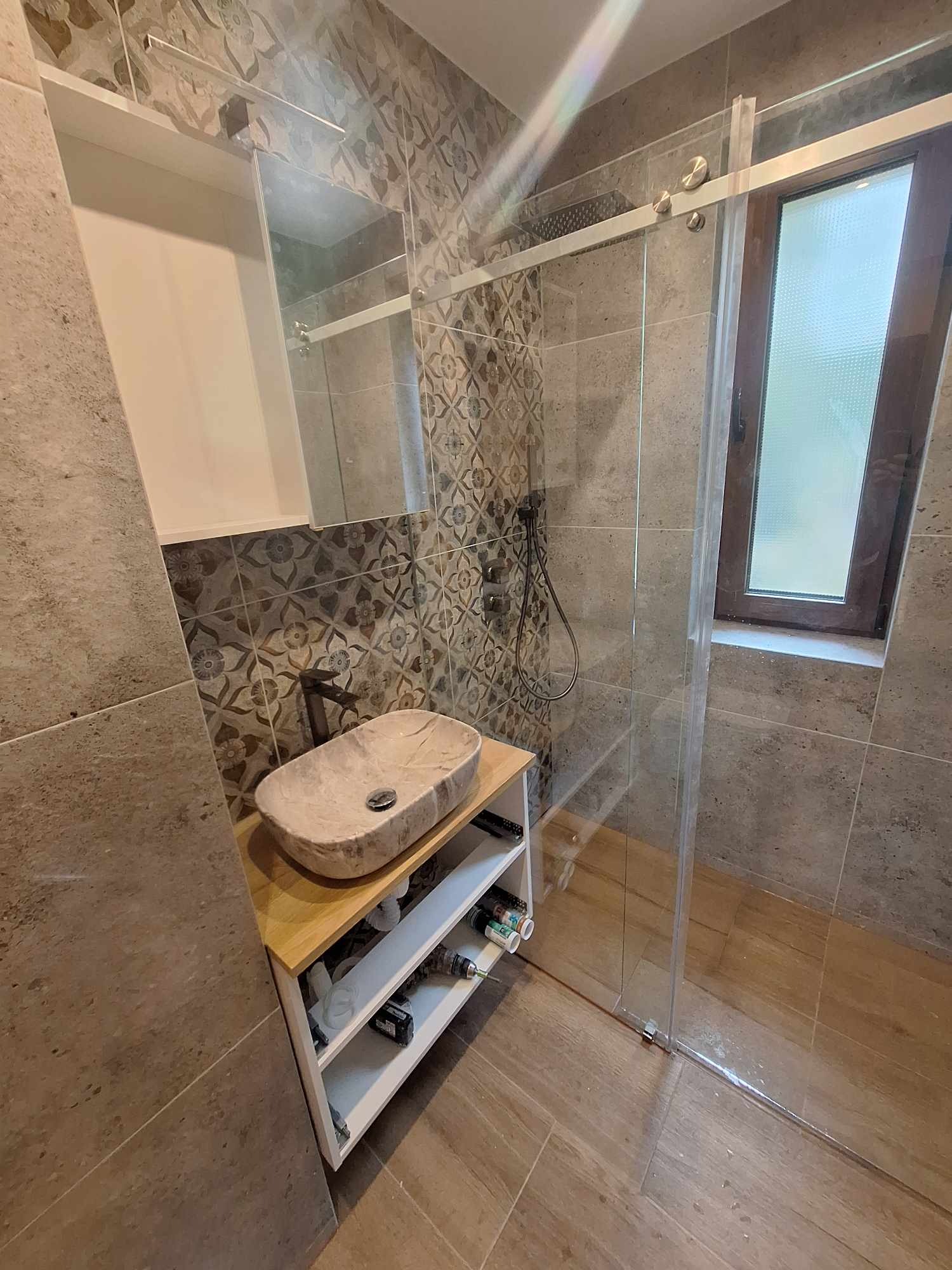 Ukończona łazienka z kamienną umywalką nablatową na drewnianym blacie, kabiną prysznicową ze szklanymi drzwiami przesuwnymi i mozaiką na ścianach.