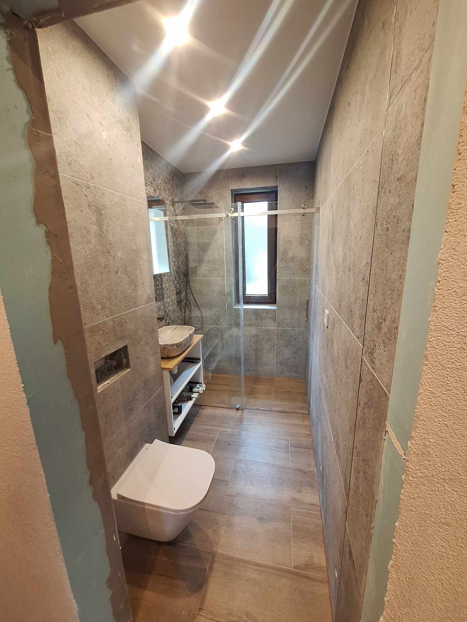 Nowoczesna łazienka w trakcie remontu, widoczne szare płytki imitujące beton, drewnopodobne panele na podłodze, biała podwieszana toaleta, umywalka nablatowa z drewnianym blatem, szklana kabina...