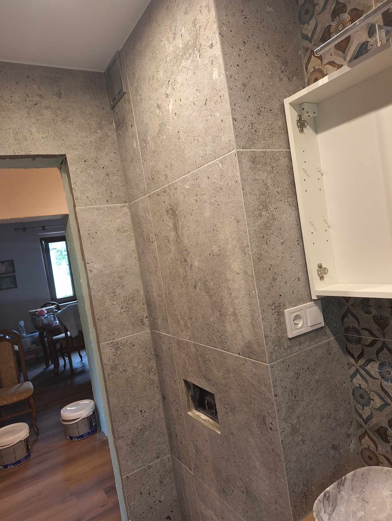 Ściana łazienki wyłożona szarymi, kwadratowymi płytkami imitującymi kamień, z widoczną białą szafką bez frontu, gniazdkiem elektrycznym i puszką instalacyjną. W tle widoczne wiaderka z zaprawą...