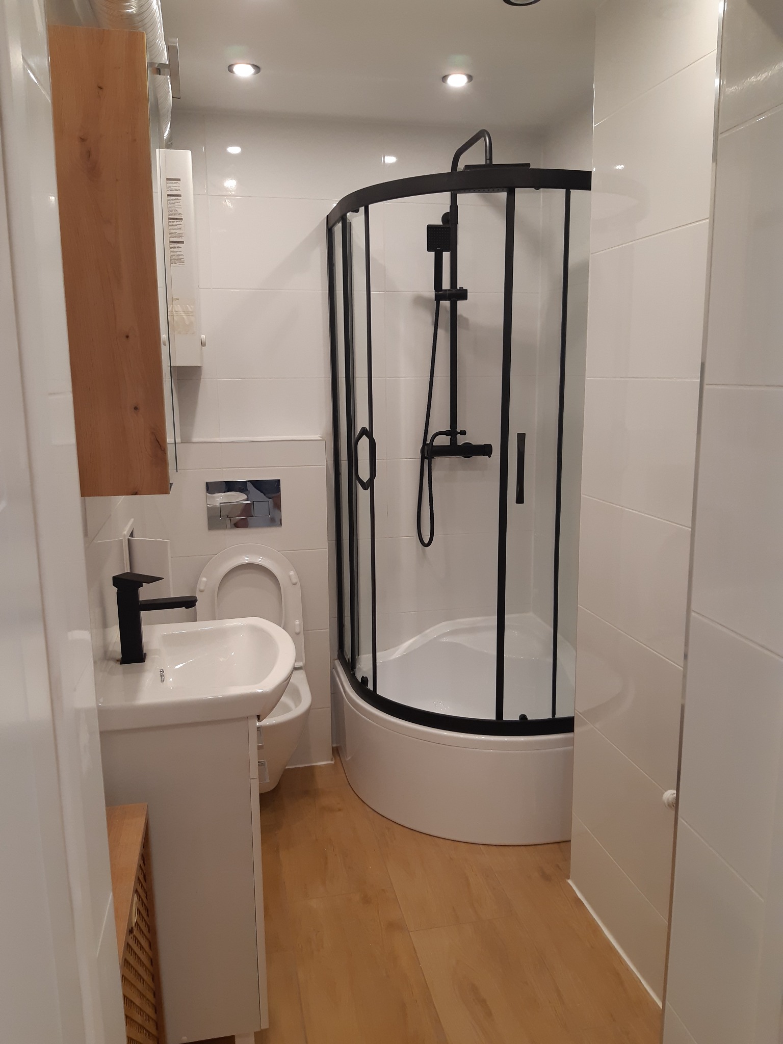 Nowoczesna łazienka z kabiną prysznicową z czarnymi profilami, białą umywalką z czarną armaturą, drewnianą szafką i białymi płytkami na ścianach oraz panelami na podłodze.