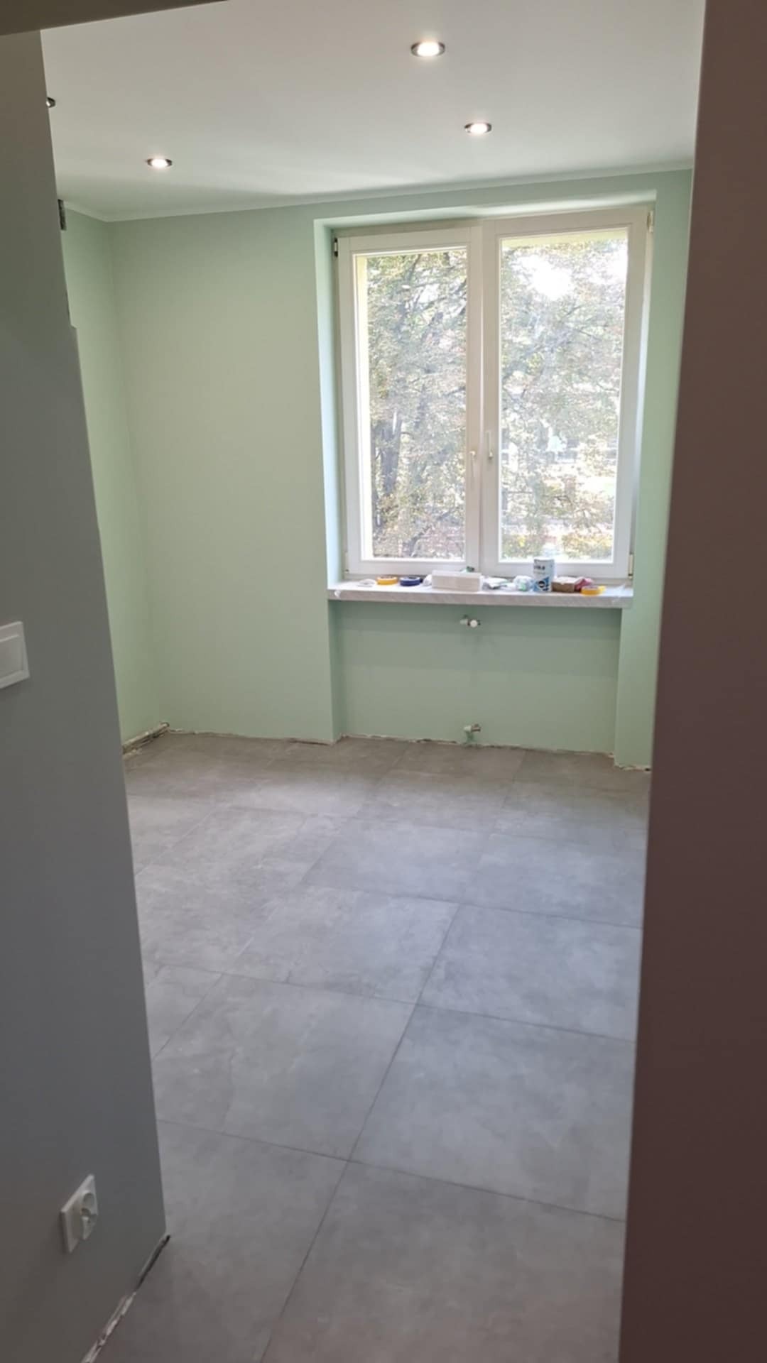 Pomalowane na jasnozielono pomieszczenie z szarymi płytkami podłogowymi, widoczne przez otwarte drzwi. Na parapecie okna stoją puszki z farbą i inne akcesoria malarskie.