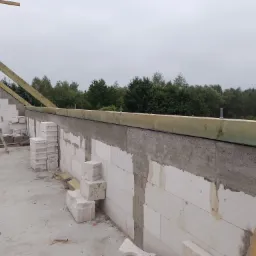 Wznoszenie ścian z białych bloczków, widoczne drewniane elementy konstrukcji dachu i drabina oparta o ścianę. W tle zalesiony teren pod pochmurnym niebem.