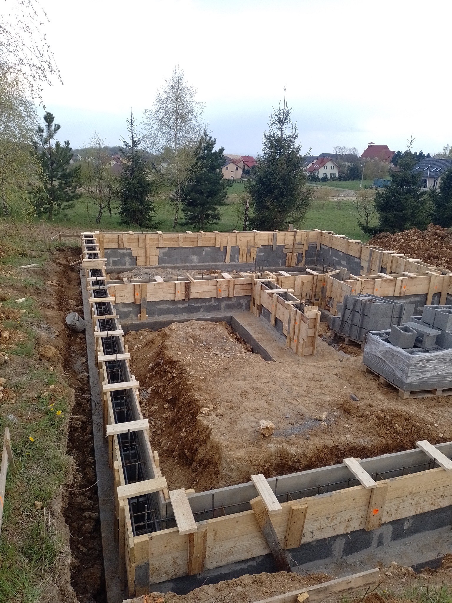 Zalane betonem zbrojenie fundamentów domu, z widocznym szalunkiem z desek i częściowo wymurowanymi ścianami fundamentowymi z bloczków betonowych, na tle krajobrazu z drzewami i zabudowaniami.