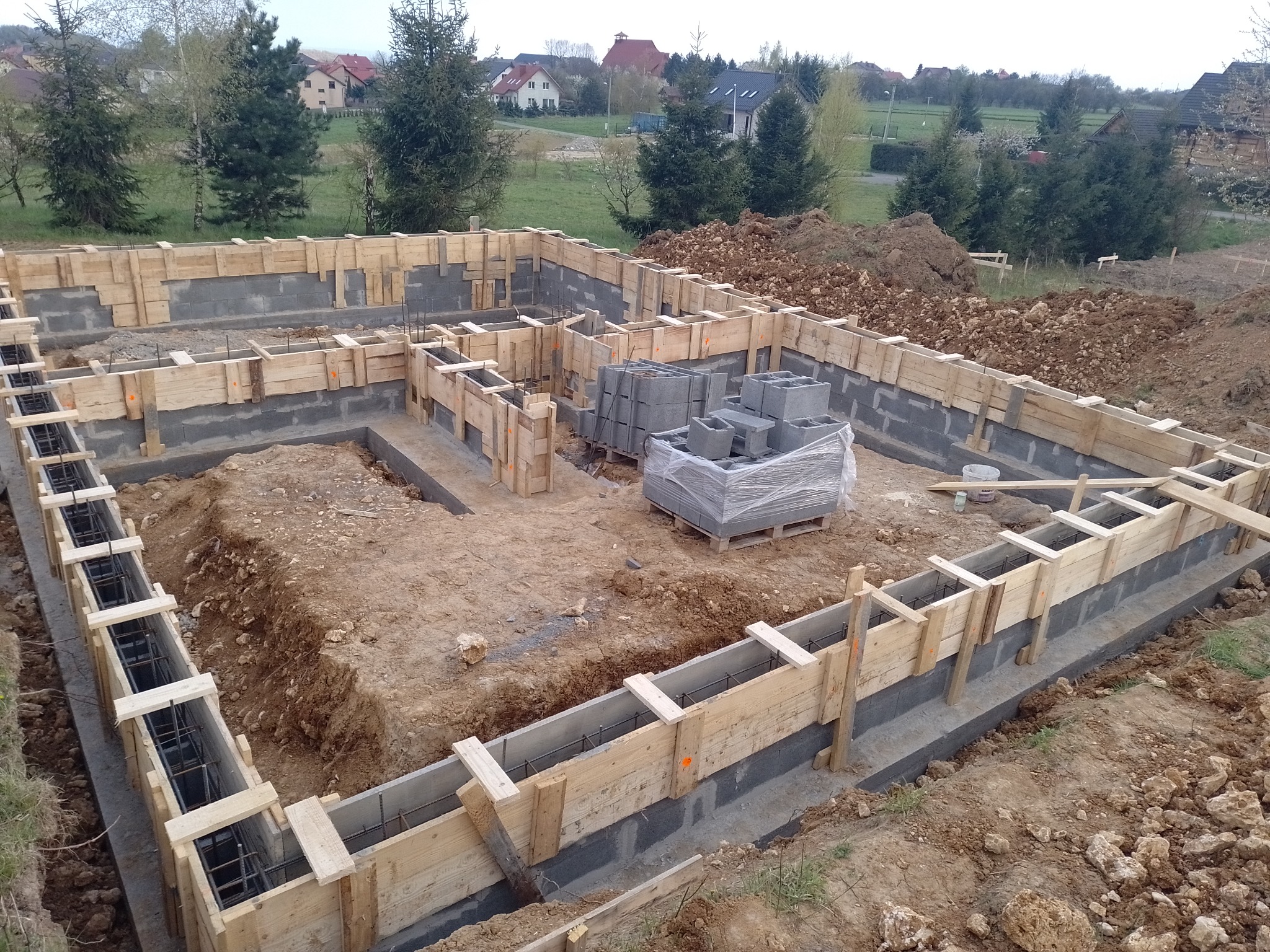 Wykopane fundamenty domu z drewnianym szalunkiem wzmocnionym stalowymi prętami zbrojeniowymi, na palecie bloczki betonowe, w tle zabudowa jednorodzinna.